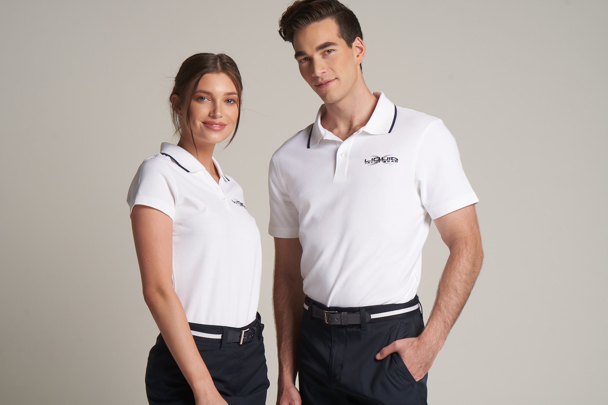 A woman and a man wearing white polos with a small logo on the left side, and dark blue shorts with a white belt.